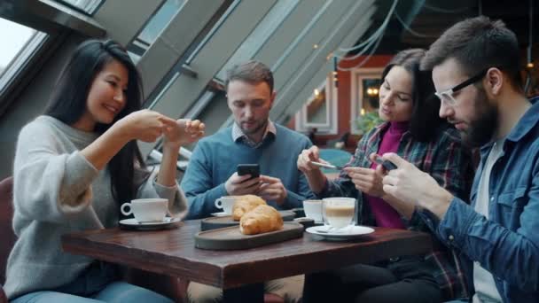 Amigos alegres tomando fotos de tazas de café y croissants en la cafetería y luego compartiendo fotos hablando — Vídeos de Stock