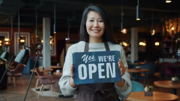 Portret pięknej azjatyckiej bizneswoman w fartuchu trzyma otwarty znak w nowoczesnej kawiarni — Wideo stockowe