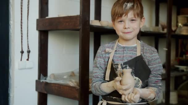 Retrato de câmera lenta de criança adorável com rosto sujo segurando vaso feito à mão na oficina de cerâmica — Vídeo de Stock