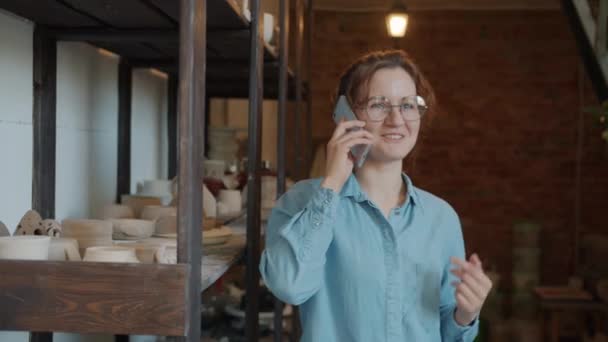 Happy young lady speaking on mobile phone in pottery workshop smiling — Stock Video