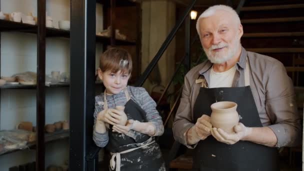 Portrait of dirty kid and senior man in aprons standing in pottery workshop holding ceramics — Stock Video