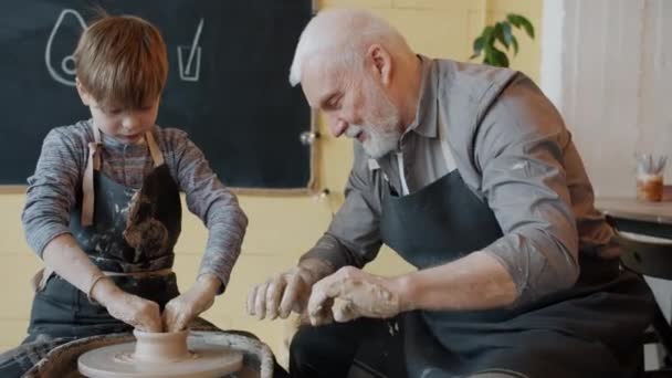 Experienced potter teaching boy to work with spinning wheel in workshop speaking — Stock Video