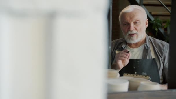 Portret van oude ambachtsman tellen keramiek in workshop voorraad en schrijven in notebook — Stockvideo