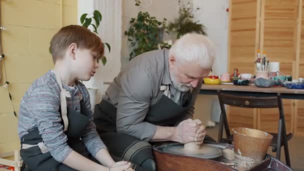 Старший гончар обучение любопытный ребенок в мастерской говорить и работать с бросать колесо — стоковое видео