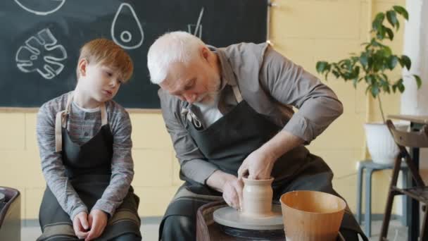 Loving grandfather making ceramic pot on throwing wheel and talking to grandchild — Stock Video