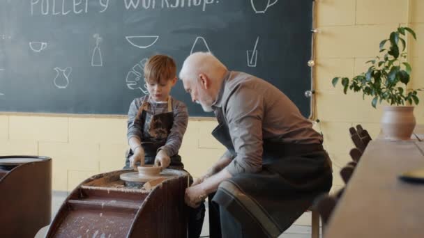 Niño creando loza utilizando la rueda de cerámica en el taller bajo la guía del hombre mayor — Vídeo de stock