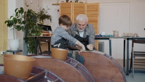 Farfar undervisar liten pojke i keramik klass med hjälp av kasta hjul arbetar med lera — Stockvideo