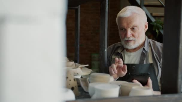 Mouvement lent de potier homme âgé en utilisant la tablette pendant l'inventaire en atelier — Video