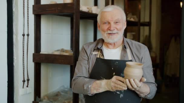 Portret van senior pottenbakker in schort staand in werkplaats met pot glimlachend — Stockvideo