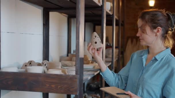 Slow motion van jonge vrouw kijken naar de hand gemaakte aardewerk vervolgens aantekeningen maken in workshop — Stockvideo