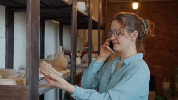 Vrolijk meisje praten op mobiele telefoon met de hand gemaakte keramiek in aardewerk studio — Stockvideo