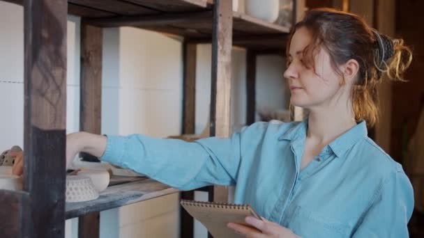 Langzame beweging van serieuze vrouwelijke pottenbakker controleren van keramiek en schrijven notities in workshop — Stockvideo