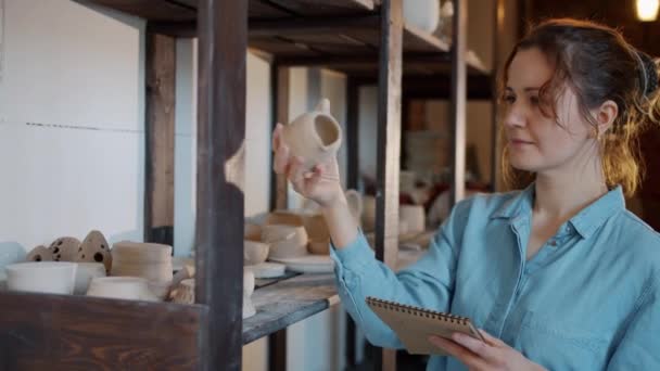 Portret van vrouwelijke pottenbakker controleren van keramiek en het schrijven van informatie in notebook in workshop — Stockvideo