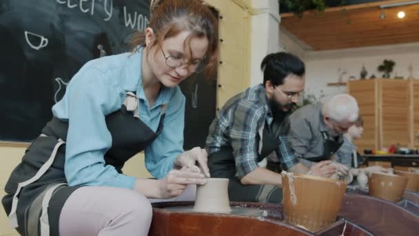 Mujer joven trabajando con arcilla en la rueda de lanzamiento durante la clase de cerámica en el taller — Vídeos de Stock