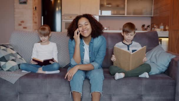 Mouvement lent de babysitter afro-américain parlant sur un téléphone portable pendant que les enfants lisent des livres à la maison — Video