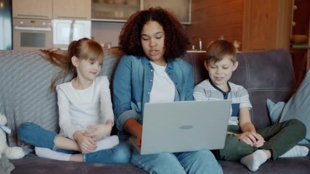 Mouvement lent des enfants espiègles s'amusant avec ordinateur portable tapant tandis que babysitter afro-américain riant — Video