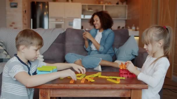 Niedliche Kinder spielen mit Holzspielzeug, während das Kindermädchen mit dem Handy telefoniert und lacht — Stockvideo