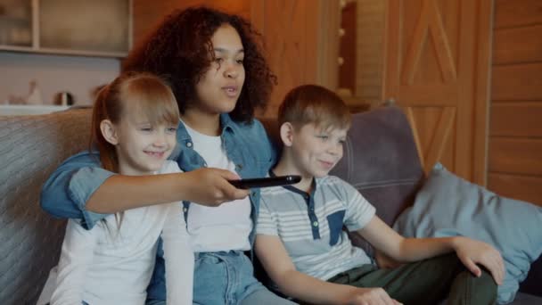 Glada kaukasiska barn tittar på TV med afroamerikansk barnflicka hemma — Stockvideo