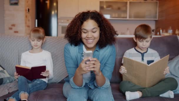 子供の男の子と女の子は自宅で本を読んでいる間に笑顔スマートフォンを使用して幸せな混合レース乳母 — ストック動画
