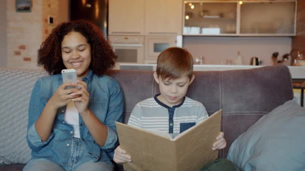 Молода жінка няня використовує смартфон під час читання дитячої книги в квартирі — стокове відео