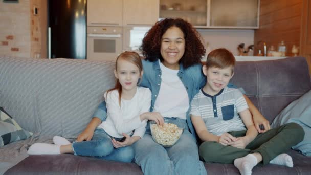 Портрет молодой афроамериканской женщины и кавказских детей, смотрящих телевизор и смеющихся дома — стоковое видео