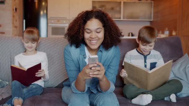 Слухняні діти читають книги, коли няня використовує смартфон і сміється вдома — стокове відео