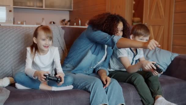 Joven niñera alegre animando a los niños jugando videojuego divertirse en el apartamento — Vídeo de stock