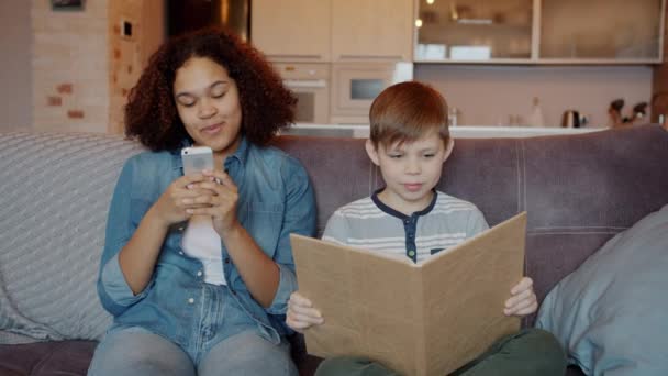 Perempuan Afro-Amerika babysitter menggunakan smartphone tersenyum saat anak laki-laki membaca buku di rumah — Stok Video