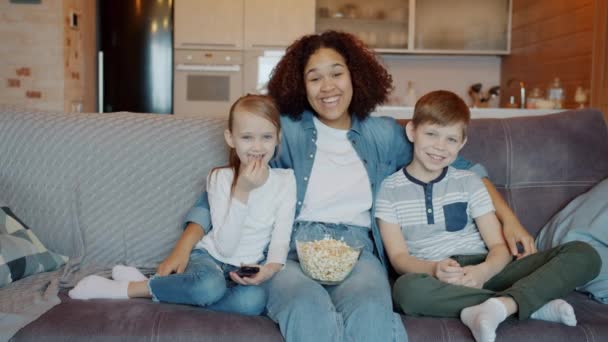 Ritratto di persone felici bambinaia e bambini che guardano la TV e ridono guardando la fotocamera a casa — Video Stock