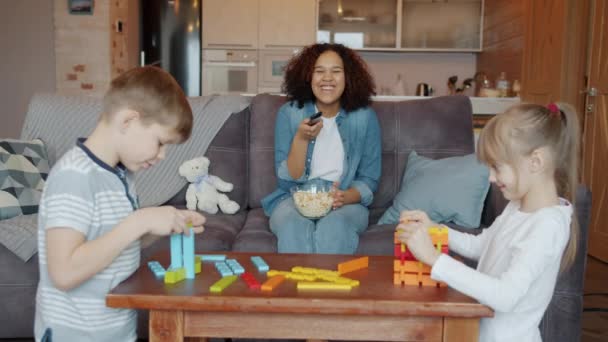 Retrato de una joven niñera viendo televisión y riendo mientras los niños juegan con juguetes — Vídeos de Stock