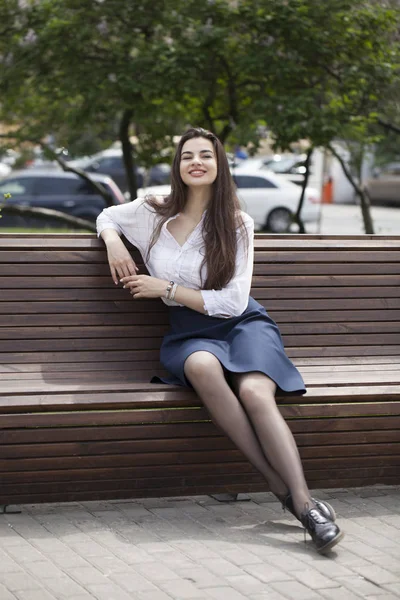Young happy brunette woman in skirt and white blouse sitting on the bench in summer park outdoors
