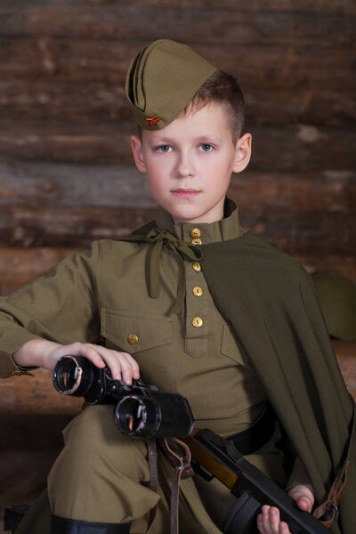 Russian boy in the old-fashioned Soviet military uniform with a binoculars on the background of a dugout from the bars