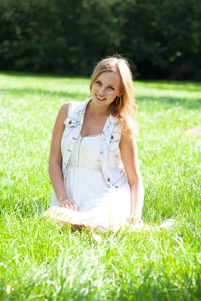 夏の公園の緑の草の上に座って白いドレスの若い美しいブロンドの女性 — ストック写真