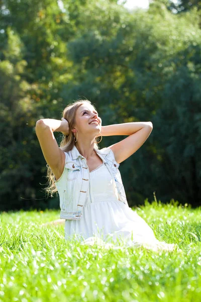 夏の公園の緑の草の上に座って白いドレスの若い美しいブロンドの女性 — ストック写真