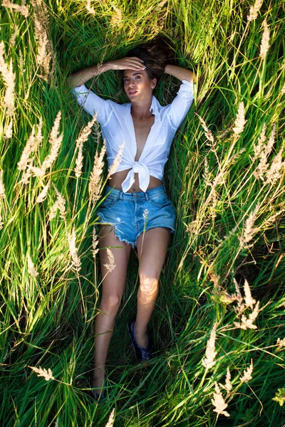 Beautiful young brunette woman in countryside on sunny day. Slim girl wearing white shirt and jeans shorts.