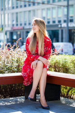Bir tam uzunlukta genç güzel sarışın kadın kırmızı yaz elbise, şehir parkta bir bankta oturur portresi