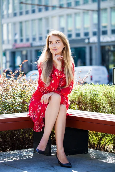 都市公園のベンチに座っている赤いサマードレスでフルレングス若い美しい金髪女性の肖像画 — ストック写真