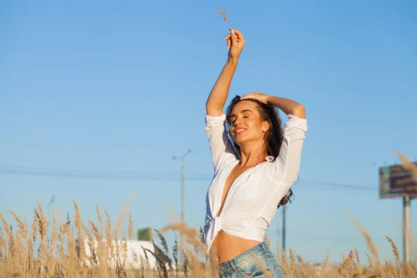 Beautiful young brunette woman in countryside on sunny day. Slim girl wearing white shirt and jeans shorts.