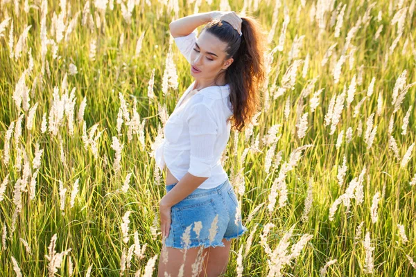 Beautiful young brunette woman in countryside on sunny day. Slim girl wearing white shirt and jeans shorts.