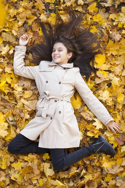 年轻美丽的小女孩在米色大衣躺在黄叶 从上面看 在秋季公园 — 图库照片