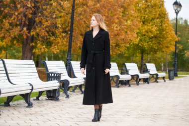 Güzel bir sarışın kız bir sonbahar park, sarı ve turuncu siyah bir ceket ile bir tam uzunlukta bırakır 