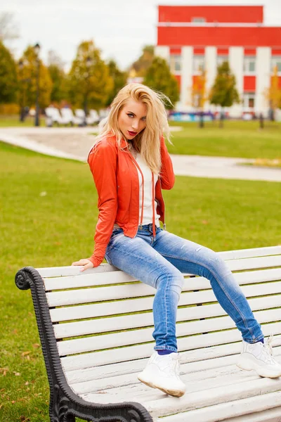 ブルー ジーンズと赤いジャケット 秋通り屋外で幸せな笑みを浮かべて美しい若い金髪女性の肖像画 — ストック写真