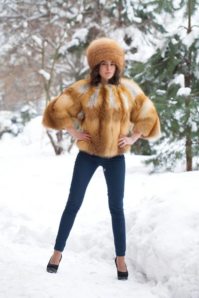 Young beautiful girl in a fur hat and a red fox fur coat posing against a winter park background