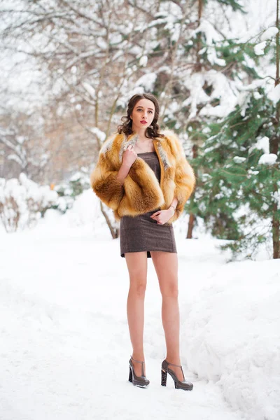 Young beautiful brunette woman in a fox fur coat posing on winter park. Model wearing stylish warm clothes.