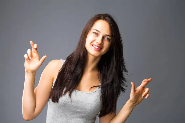 Body language and emotions concept. Glad dark haired beautiful satisfied girl beggs God to help her, keeps fingers crossed, wishes good luck, prays indoor isolated over gray studio background.