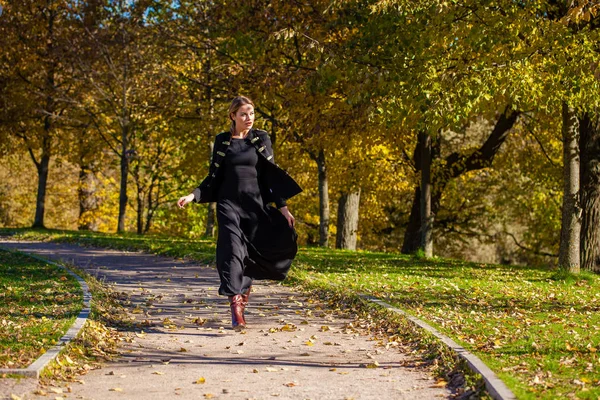 秋の公園を歩く黒のドレスで美しい若いブロンドの女性 — ストック写真