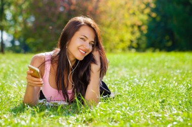 Elbiseli Genç güzel kadın yeşil çimenlerin üzerinde yatıyor ve akıllı telefon kulaklık müzik dinler.