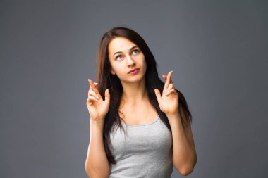 Beden dili ve duygular kavramı. Memnun koyu saçlı güzel memnun kız beggs Tanrı ona yardım için parmak çarpı işareti tutar, iyi şanslar diliyor, gri studio arka plan üzerinde kapalı izole dua ediyor.