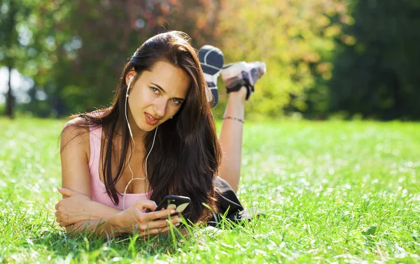 Young woman in dress lies on green grass and listens to music in — Stock Photo, Image