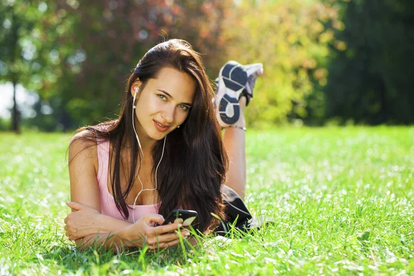 Young woman in dress lies on green grass and listens to music in — Stock Photo, Image